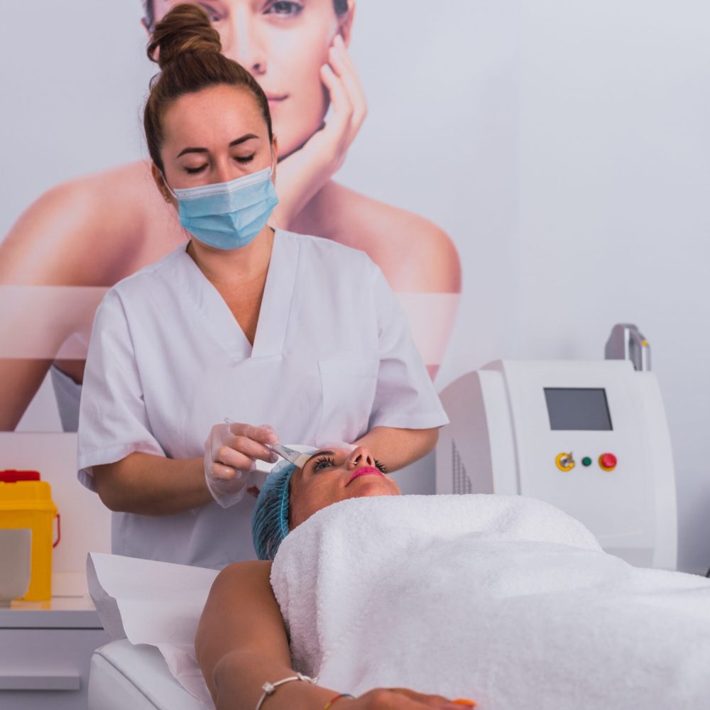 Front view of a cosmetologist making a beauty treatment applying vitamins and gold mask to face of adult woman for rejuvenation in the spa center.